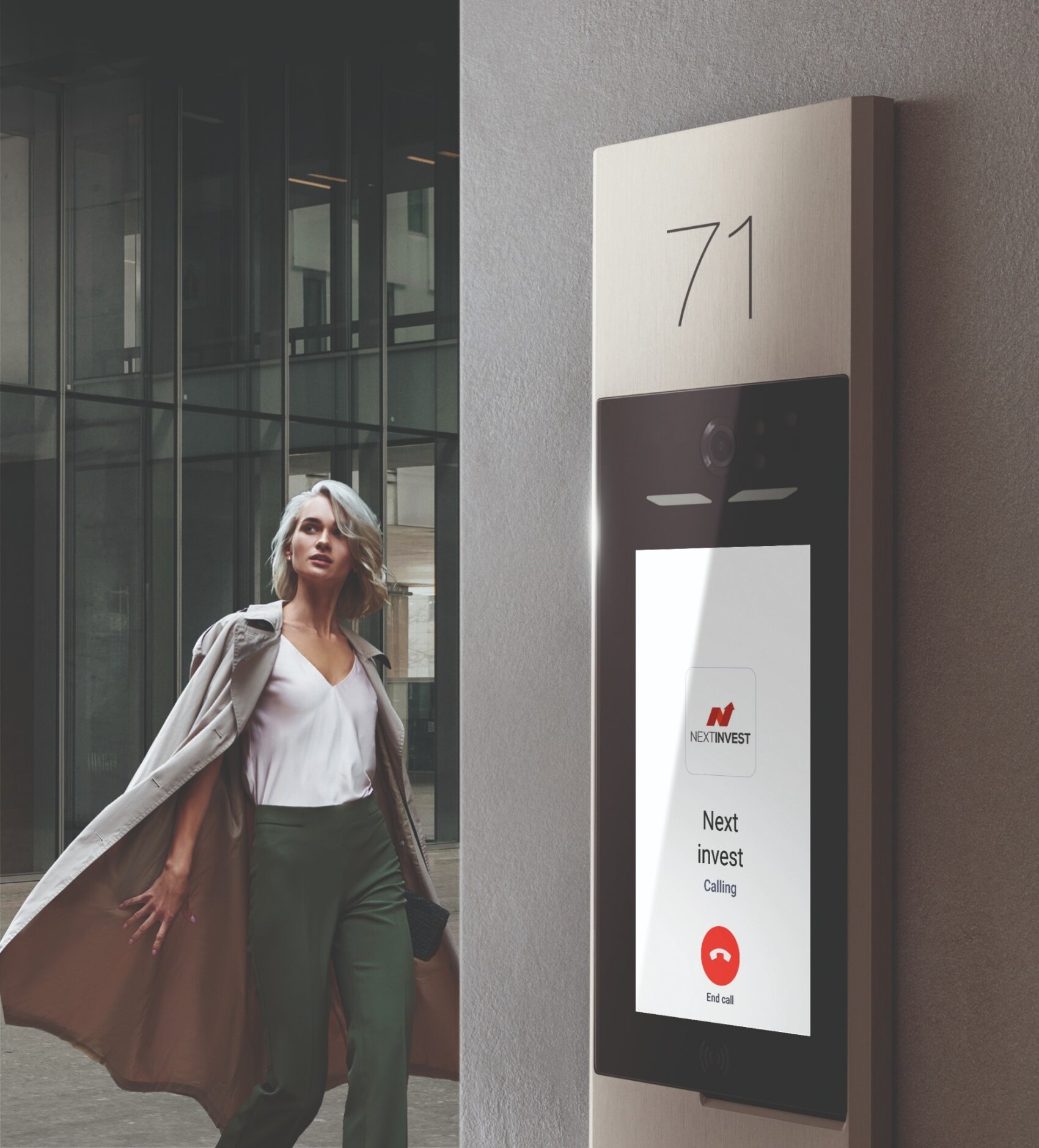 Une femme aux cheveux argentés et en trench-coat marche près de l'entrée d'un bâtiment doté d'un interphone à écran tactile affichant Next Invest Calling et un bouton de fin d'appel rouge sous le numéro 71.